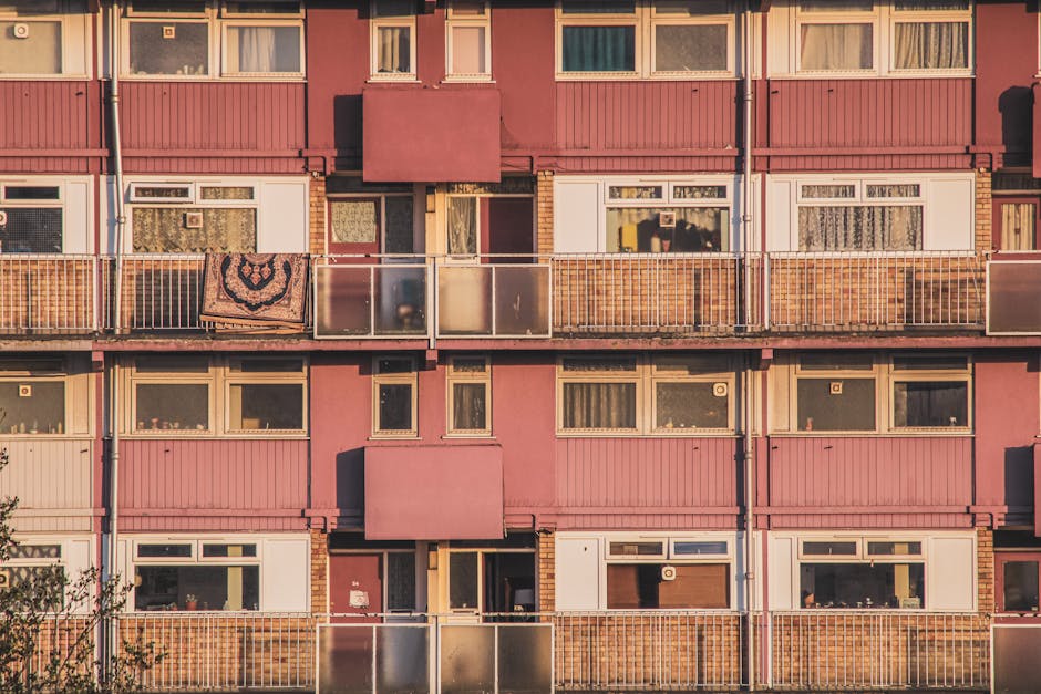 A close-up view of the exterior façade of a multistory residential building with balconies on each floor. The building features pink panels and white window frames, with some balconies enclosed with g